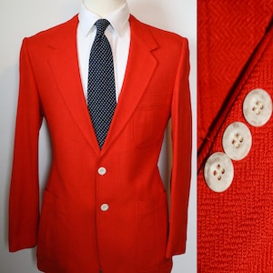 May include: A bright red blazer with two white buttons and a notched lapel. The blazer is paired with a white dress shirt and a navy blue tie with white polka dots. The blazer has two front pockets.