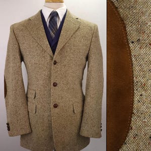 May include: A tan tweed blazer with brown elbow patches and brown buttons. The blazer has a notched lapel, two front pockets, and a three-button closure. A blue vest and striped tie are visible underneath.