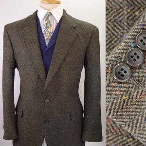 May include: A brown herringbone tweed blazer with multicolored flecks, featuring a two-button front closure and flap pockets. The blazer is layered over a blue vest, a white collared shirt, and a floral patterned necktie. Three buttons are on the sleeve.