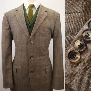 May include: A brown herringbone tweed blazer with a subtle windowpane check pattern, featuring three front buttons, two flap pockets, and a notched lapel. The blazer is worn over a white collared shirt, a green textured vest, and a green necktie.