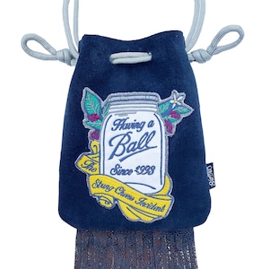 May include: A navy blue suede pouch with a drawstring closure and a decorative fringe. The pouch features an embroidered patch with a mason jar design and the text "Having a Ball Since 1998" and "The Stung Cheese Incident".