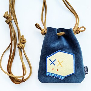 May include: A small blue velvet drawstring bag with a tan leather cord. The bag has a white and yellow embroidered patch with a geometric design and two red dots. The patch has a blue outline and a row of blue triangles along the bottom.