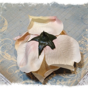 May include: An artificial flower head featuring soft, pale cream-colored petals with subtle pink-tinted edges. The center of the flower is anchored by a dark green, star-shaped plastic calyx with a small, protruding stem attachment point at the top.