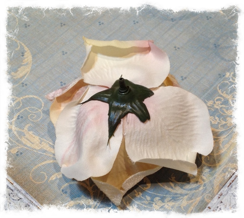 May include: An artificial flower head featuring soft, pale cream-colored petals with subtle pink-tinted edges. The center of the flower is anchored by a dark green, star-shaped plastic calyx with a small, protruding stem attachment point at the top.