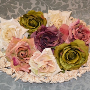 May include: An oval decorative tray with a raised floral border holds a collection of seven artificial roses in various colors, including cream, olive green, dusty pink, and deep plum, featuring layered fabric petals and realistic textures.