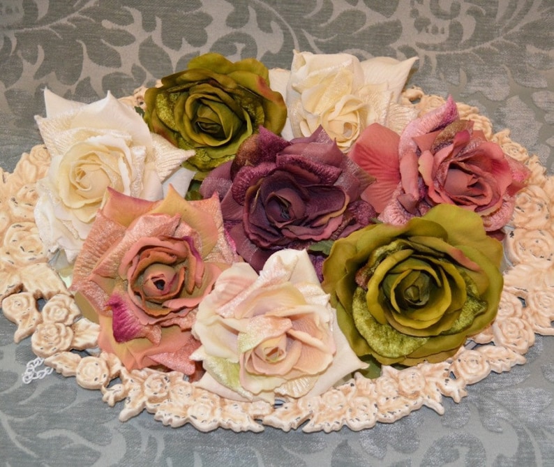 May include: An oval decorative tray with a raised floral border holds a collection of seven artificial roses in various colors, including cream, olive green, dusty pink, and deep plum, featuring layered fabric petals and realistic textures.