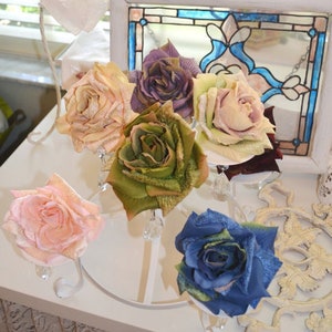 May include: A collection of six artificial roses in various colors including peach, purple, light green, pink, olive green, and blue, each featuring textured petals with a subtle glitter finish, displayed on a white metal stand with crystal accents.