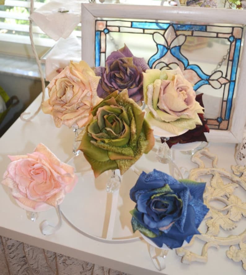 May include: A collection of six artificial roses in various colors including peach, purple, light green, pink, olive green, and blue, each featuring textured petals with a subtle glitter finish, displayed on a white metal stand with crystal accents.