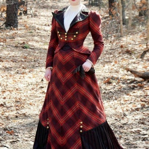 May include: A woman wearing a red and black plaid dress with a black hat and gloves. The dress has a fitted bodice and a full skirt with a black trim. The woman is standing in a wooded area.