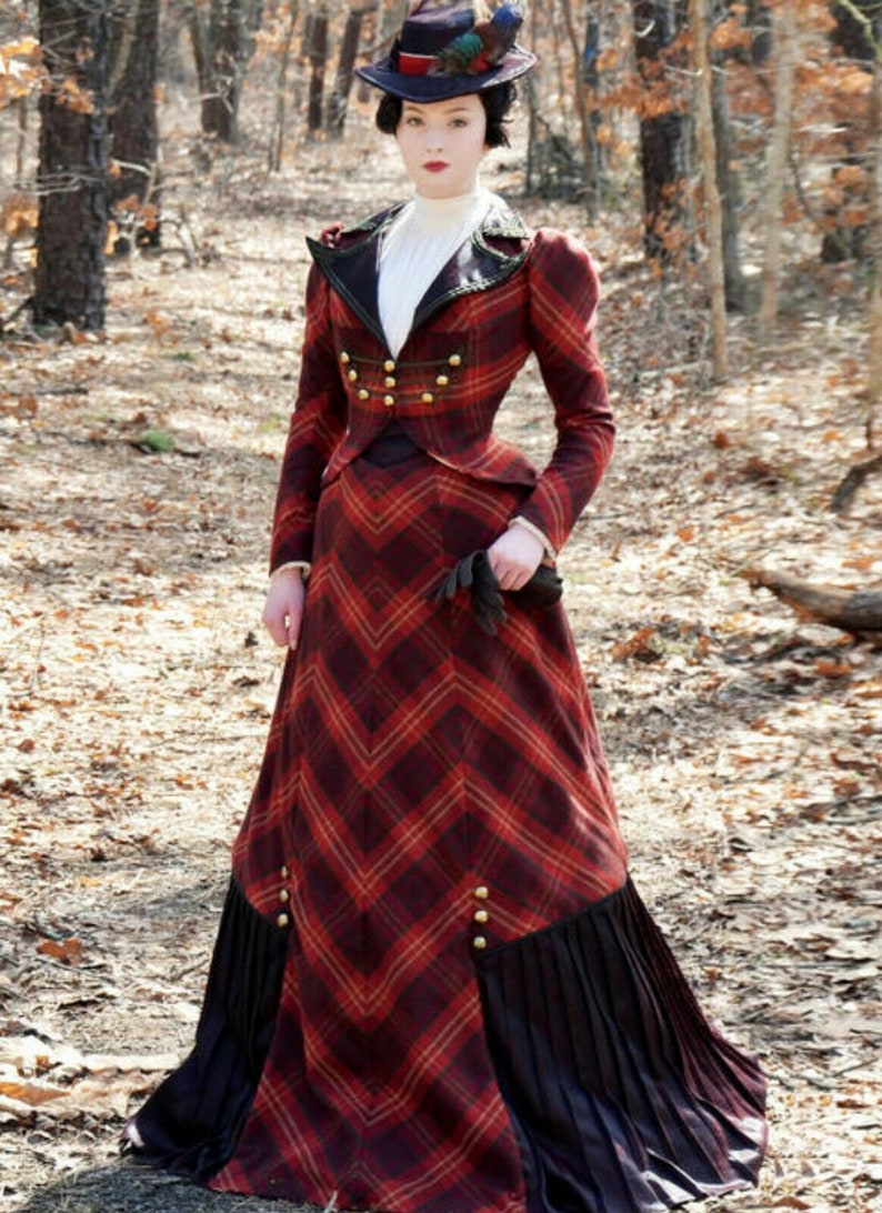 May include: A woman wearing a red and black plaid dress with a black hat and gloves. The dress has a fitted bodice and a full skirt with a black trim. The woman is standing in a wooded area.