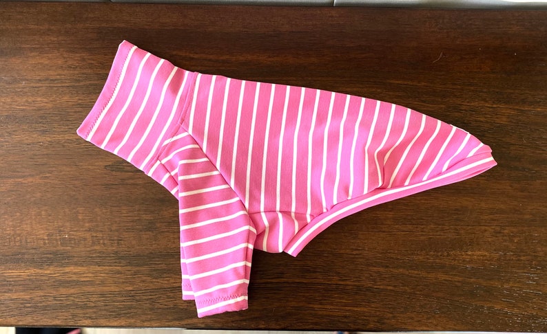 May include: Pink and white striped dog shirt. The shirt has a turtleneck collar and is made of a soft, stretchy fabric.