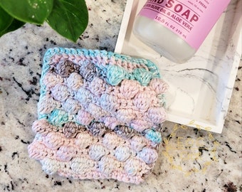 Könnte beinhalten: Ein gehäkelter Geschirrtuch mit einem strukturierten Blasendesign in Rosa-, Blau- und Grautönen. Das Geschirrtuch liegt auf einer weißen Marmorarbeitsplatte neben einer Flasche Meyer's Clean Day Handseife mit Pfingstrosenduft.