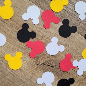 May include: Scattered paper cutouts in the shape of Mickey Mouse's head in yellow, red, white, and black. The cutouts are scattered on a light brown wooden surface.