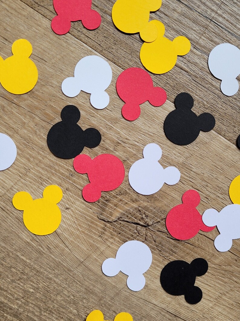 May include: Scattered paper cutouts in the shape of Mickey Mouse's head in yellow, red, white, and black. The cutouts are scattered on a light brown wooden surface.
