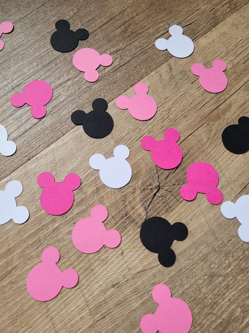 May include: Scattered confetti in the shape of a mouse head in black, white, and pink. The confetti is scattered on a wooden surface. The confetti is a party decoration.