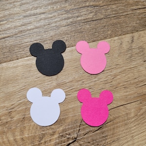 May include: Four die-cut Mickey Mouse head shapes in black, pink, white, and magenta. The shapes are arranged on a wooden surface, with the black shape in the upper left, pink in the upper right, white in the lower left, and magenta in the lower right.