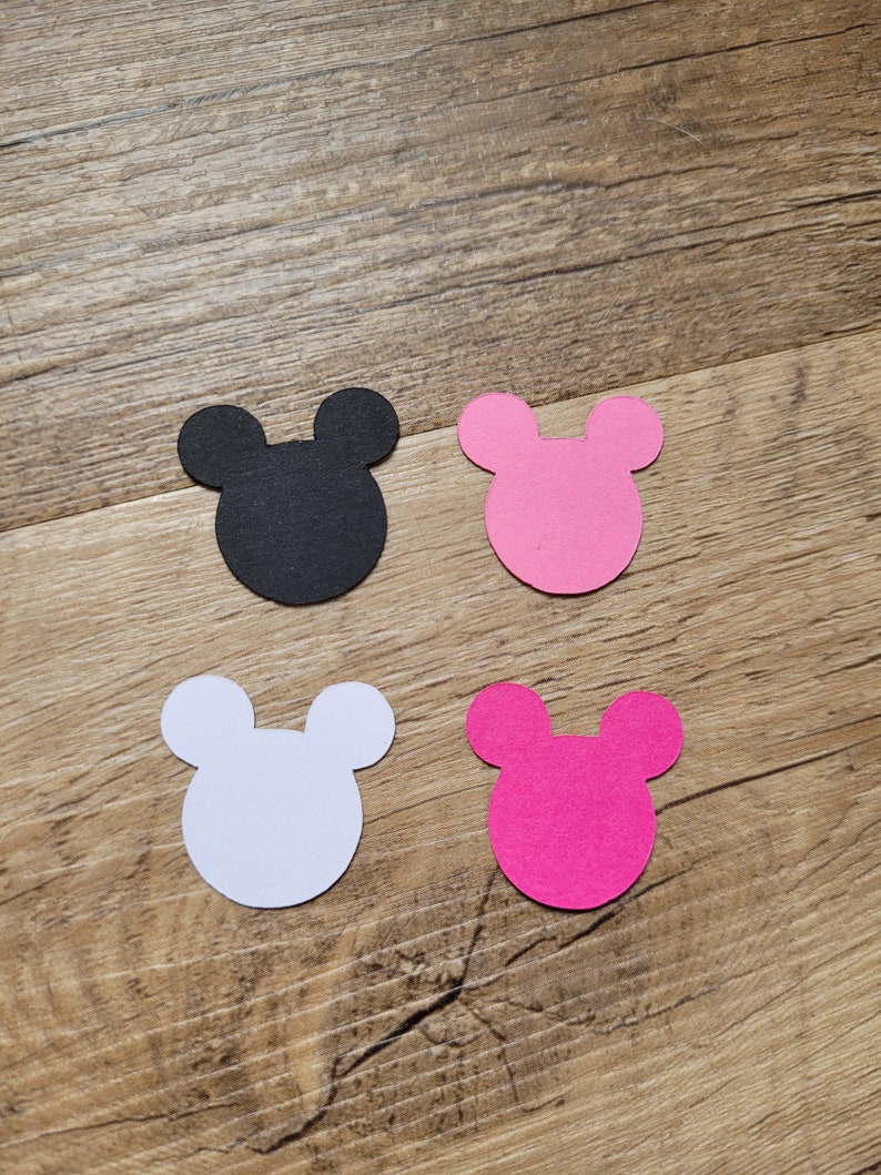 May include: Four die-cut Mickey Mouse head shapes in black, pink, white, and magenta. The shapes are arranged on a wooden surface, with the black shape in the upper left, pink in the upper right, white in the lower left, and magenta in the lower right.