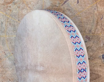 May include: A round, light brown frame drum with a light blue, pink, and orange patterned border. The drum is made of wood and has a natural leather head.