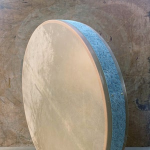 Op de afbeelding: Een grote, ronde, lichtbruine raamtrommel met een blauwe, gepatroneerde rand. De trommel is gemaakt van hout en heeft een natuurlijk leren vel.