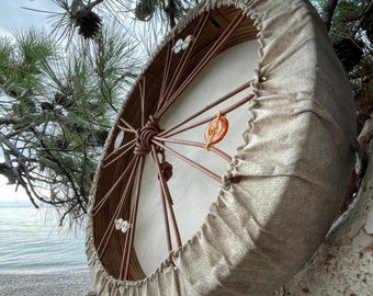May include: A round, handmade drum with a light tan fabric cover and a natural wood frame. The drum is decorated with small, clear and orange beads. The drum is hanging from a tree branch near a beach.