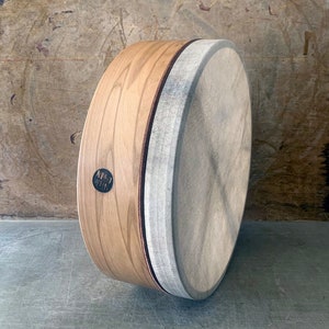 May include: A wooden frame drum with a natural colored head. The drum is made of light brown wood with a dark brown rim. The head is made of a natural colored material and has a slightly textured surface. The drum is labeled "KIED WOODS" in black lettering.