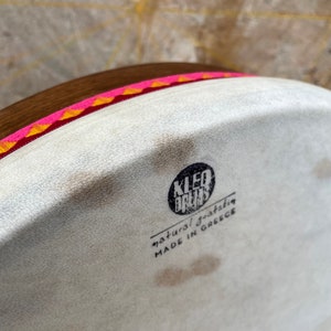 May include: A white drum with a brown wooden frame and a pink and orange trim. The drum has a black label that reads "KLEO DRUMS natural goatskin MADE IN GREECE".