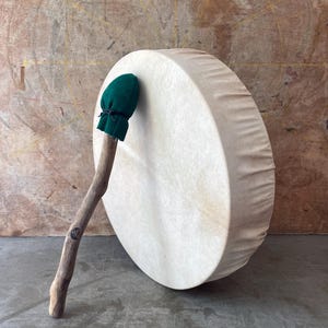 May include: A large, round drum with a natural hide surface. A wooden drumstick with a green fabric head rests against the drum. The drum and stick are set against a neutral, textured background.