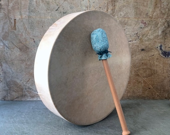 Può includere: Un grande tamburo rotondo con una pelle color beige chiaro e una mazza di legno. La mazza ha una testa a fantasia blu chiaro e un lungo manico in legno naturale. Il tamburo è appoggiato su uno sfondo strutturato dai toni neutri.