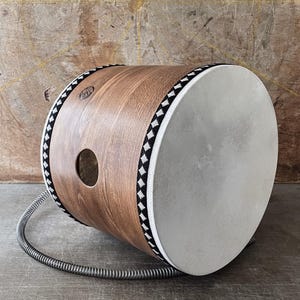 May include: A cylindrical wooden drum with a light brown finish. The drumhead is white, and the edges are trimmed with a black and white geometric pattern. The drum is supported by a coiled metal stand. The drum's diameter is approximately 30 cm.