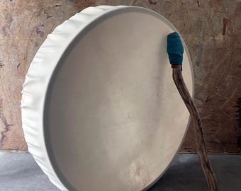May include: A round frame drum with a smooth, off-white drumhead stretched over the frame. A wooden mallet with a teal-colored fabric-wrapped head rests against the side of the drum, featuring a natural, textured branch handle.