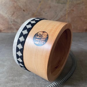 Puede incluir: Un casco de tambor cilíndrico de madera con acabado en tono claro, que presenta un borde de tela con patrón de diamantes en blanco y negro. Una pegatina circular con el texto KLEO DRUMS está adherida al lateral, junto a un muelle metálico.