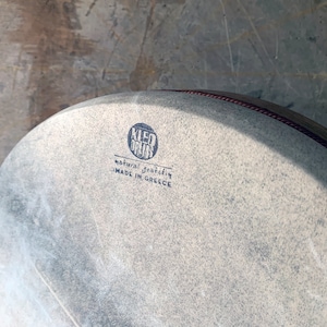 May include: A close-up of a light gray drum head with a dark blue logo that reads "KLED DRUMS" and the text "natural goat skin MADE IN GREECE".