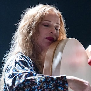 May include: A woman with long, wavy blonde hair, wearing a patterned top, embraces a large, round drum. Her eyes are closed, and she holds a red mallet, suggesting a musical performance. The drum has a light-colored surface and a wooden frame.