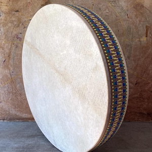 Op de afbeelding: Een ronde frametrommel met een glad, gebroken wit dierenvel. De houten rand is omwikkeld met een decoratieve geweven band met een herhalend geometrisch patroon in blauwe, gele en groene tinten, wat zorgt voor een textuurrijke en kleurrijke rand.