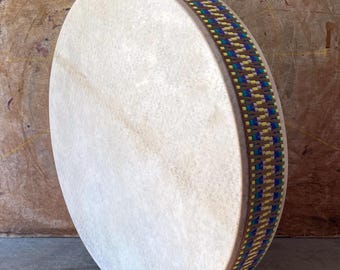 May include: A circular frame drum with a smooth, off-white animal skin head. The wooden shell is wrapped in a decorative woven band featuring a repeating geometric pattern in shades of blue, yellow, and green, creating a textured, colorful border.