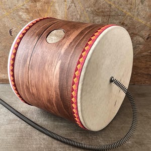 Puede incluir: Un tambor artesanal con acabado de veta de madera, un parche de color claro y un ribete decorativo rojo, amarillo y naranja. Un resorte metálico se extiende desde el tambor. El tambor tiene un orificio de sonido circular.