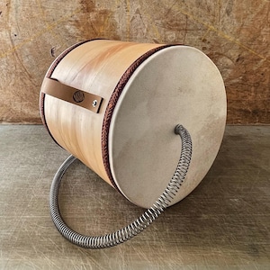 Puede incluir: Un tambor cilíndrico de madera clara con un acabado marrón claro y un asa de cuero beige. El tambor tiene un borde marrón y rojo y un resorte metálico que se extiende desde un extremo. El tambor está ligeramente inclinado.
