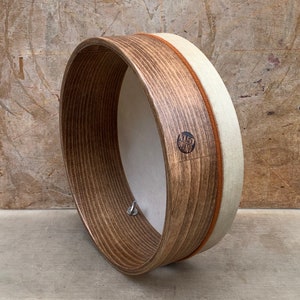 Può includere: Un tamburo a cornice in legno marrone con una pelle bianca e una cinghia in pelle beige. Il tamburo ha una chiave di accordatura e un logo che dice "KLEO DRUMS".