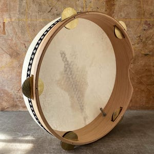 May include: A wooden frame drum with a white head and black and white trim. The drum has four brass jingles on the side.