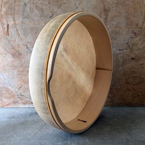 May include: A wooden frame drum with a natural leather head and a gold trim around the edge. The drum is resting on a gray surface.