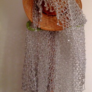 Silver lurex shawl folded over a straw hat on the stand, showing the airy crochet pattern and delicate bead trim.