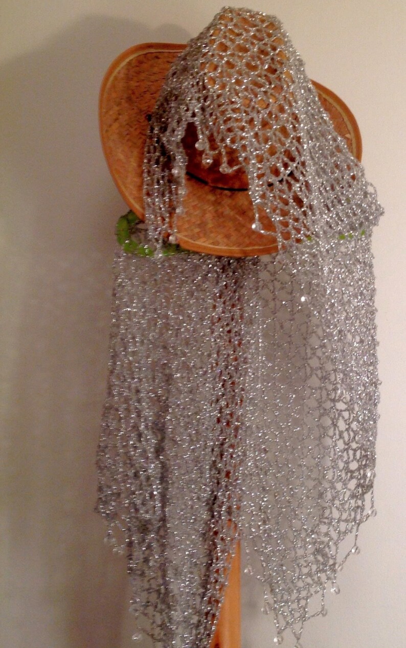 Silver lurex shawl folded over a straw hat on the stand, showing the airy crochet pattern and delicate bead trim.