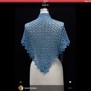 May include: A light blue triangular shawl featuring an intricate openwork crochet pattern with a scalloped edge. The garment is draped over a mannequin, showcasing the detailed yarn texture and the repeating geometric stitch design throughout.