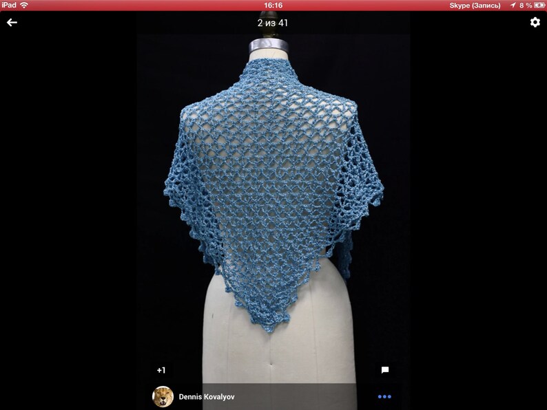 May include: A light blue triangular shawl featuring an intricate openwork crochet pattern with a scalloped edge. The garment is draped over a mannequin, showcasing the detailed yarn texture and the repeating geometric stitch design throughout.