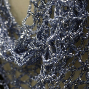 May include: A light blue scarf featuring an open, loose-knit crochet pattern. The yarn has a metallic, shimmering quality that catches the light, creating a textured, airy appearance with visible interconnected loops throughout the fabric.