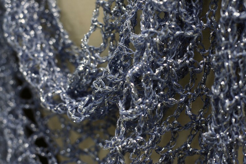 May include: A light blue scarf featuring an open, loose-knit crochet pattern. The yarn has a metallic, shimmering quality that catches the light, creating a textured, airy appearance with visible interconnected loops throughout the fabric.