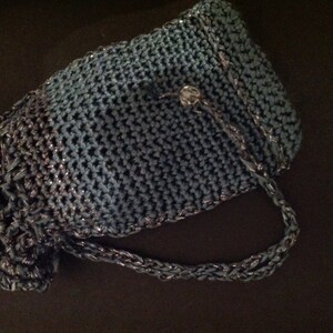 Grey crochet clutch on a dark surface, side view showing the woven strap and button detail.