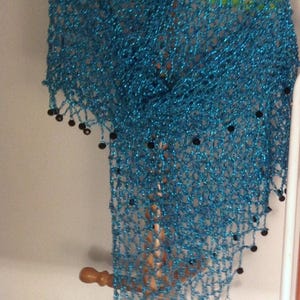 Turquoise lurex crochet shawl hanging beneath a straw hat on a wooden stand, its long shimmering drape edged with dark beads.