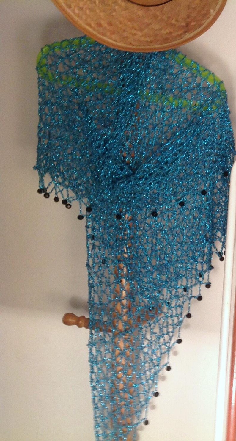 Turquoise lurex crochet shawl hanging beneath a straw hat on a wooden stand, its long shimmering drape edged with dark beads.