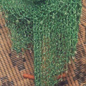 Green lurex crochet shawl hanging on a wooden stand beside a woven panel, full-length view of the shimmering wrap.